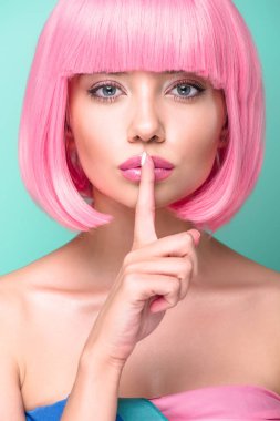 close-up portrait of young woman with pink bob cut showing silence gesture and looking at camera isolated on turquoise clipart