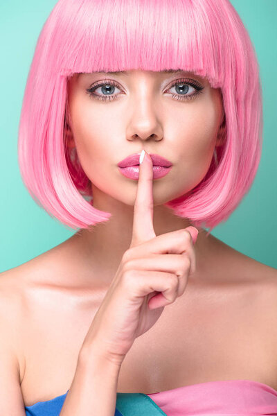 close-up portrait of young woman with pink bob cut showing silence gesture and looking at camera isolated on turquoise