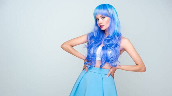 serious young woman with bright blue hair and skirt standing with arms akimbo and looking at camera isolated on grey