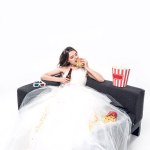 Hungry young bride in wedding dress sitting on couch with beer and junk food on white