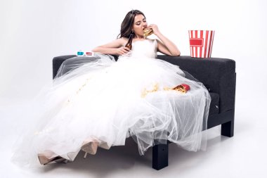 kanepede oturan ve bira ile burger yemek üzerine beyaz gelinlikle depresif aç genç gelin