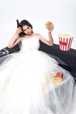 depressed young bride in wedding dress sitting on couch with beer and junk food on white clipart