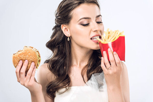 young bride in wedding dress eating burger and french fries isolated on white