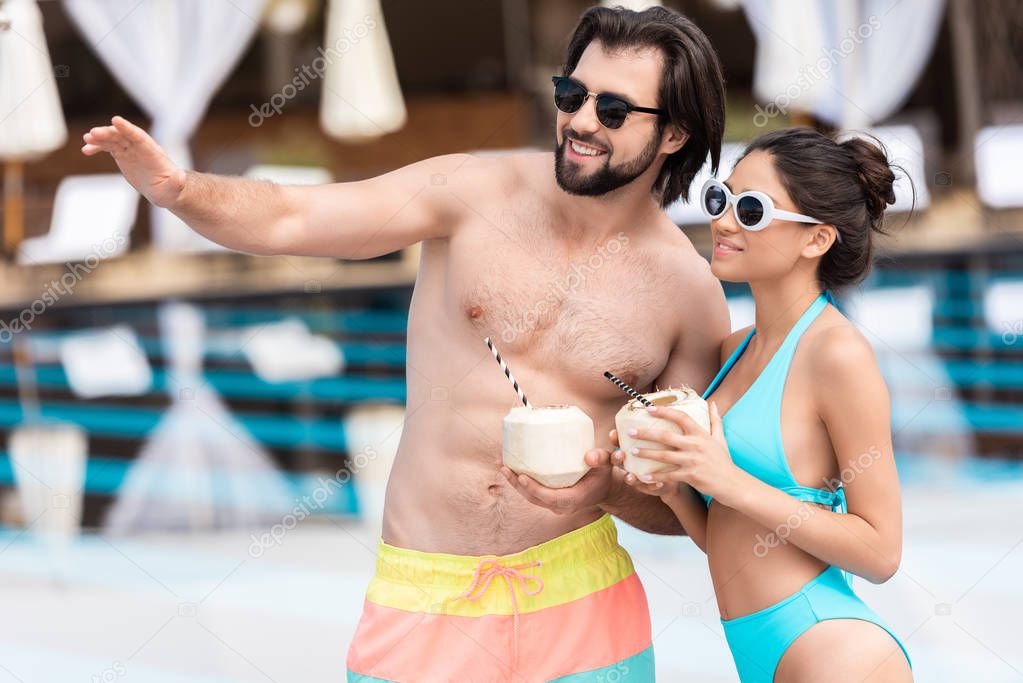 young couple in sunglasses with coconut cocktails, man showing something