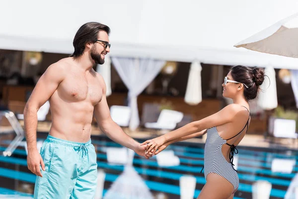スイミング プールのそばで手を繋いでいるサングラスで幸せな美しいカップル  — 無料ストックフォト