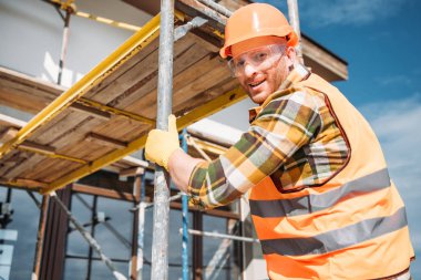 handsome builder climbing on scaffolding at construction site and looking at camera clipart