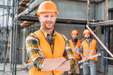 smiling builder standing at construction site with crossed arms while his colleagues standing blurred on background