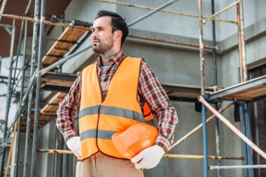 handsome builder in reflective vest holding helmet while standing in front of building house and looking away clipart