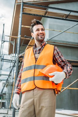 happy builder in reflective vest holding helmet while standing in front of building house and looking away clipart