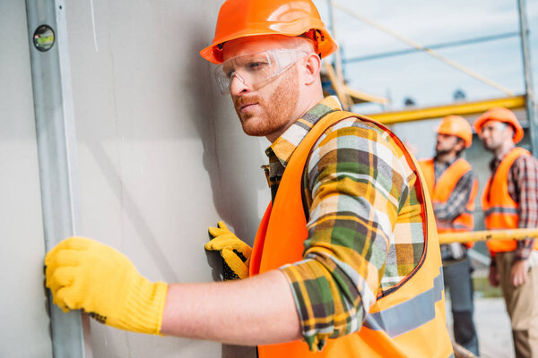 concentrated builder using bubble level at construction site while his colleagues standing on background