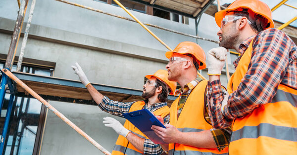 wide shot of group of builders in hard hats and reflective vests pointing at building house