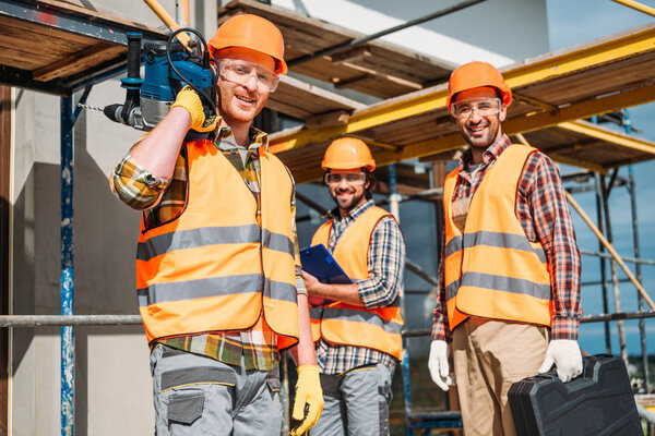 group of smiling builders with building equipment standing at construction site and looking at camera