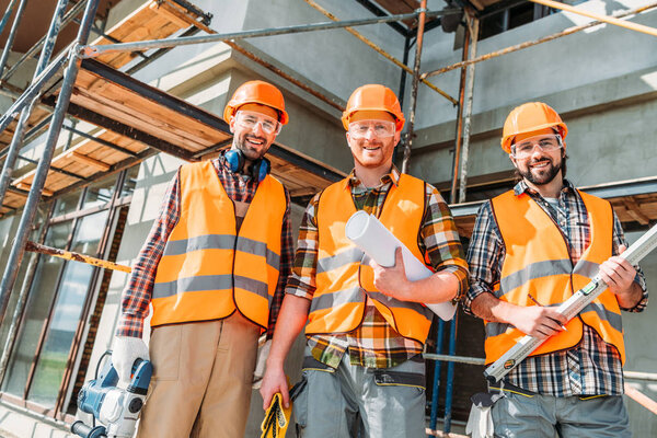 вид снизу группы счастливых оборудованных строителей, смотрящих на камеру на строительной площадке
