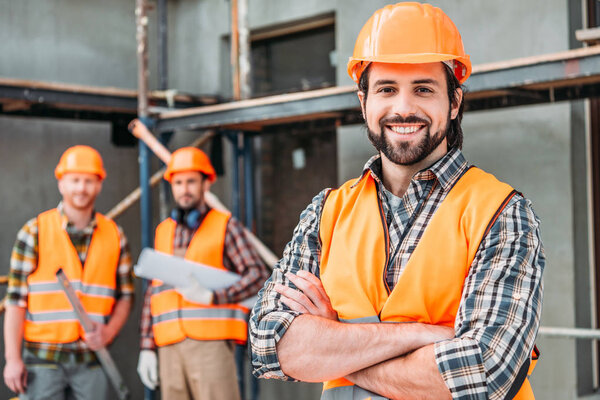 красивый улыбающийся строитель стоит на стройке со скрещенными руками, в то время как его коллеги стоят размытые на заднем плане
