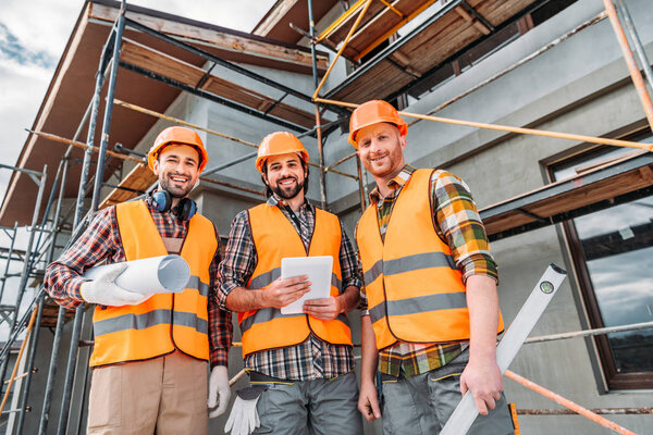 bottom view of group of smiling builders with blueprint and tablet looking at camera at construction site