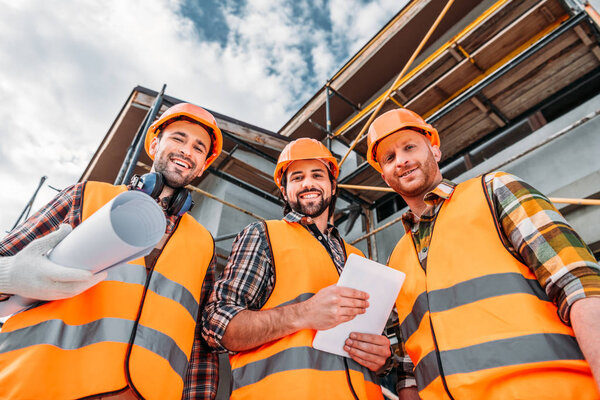 bottom view of group of builders with blueprint and tablet looking at camera at construction site