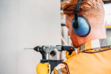 confident builder in noise reducing headphones using power drill at construction site clipart