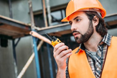 close-up portrait of handsome builder in protective helmet and vest using walkie talkie at construction site clipart