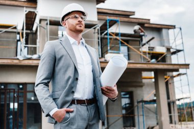 handsome architect in suit and hard hat holding blueprint standing in front of building house and looking away clipart