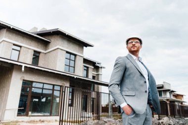 handsome architect in suit and hard hat standing in front of building house clipart