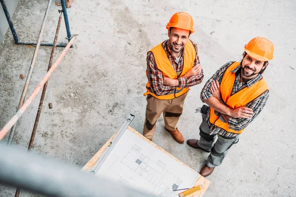 高角度的微笑建设者与蓝图站在建筑工地 看着相机 — 图库照片