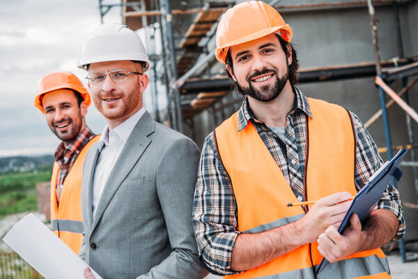 строители и архитектор с чертежом и буфером обмена, смотрящие в камеру
