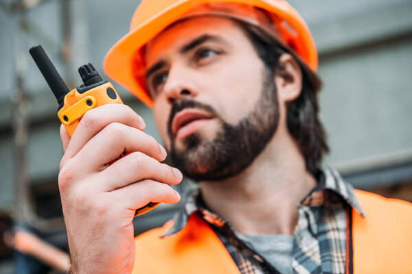 close-up portrait of builder in protective helmet and vest using walkie talkie at construction site