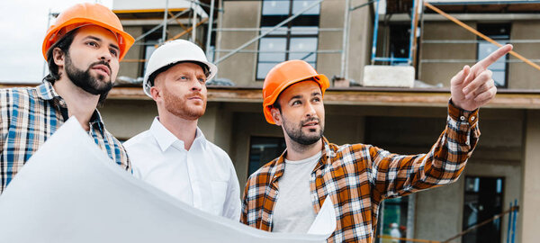 wide shot of group of architects with building plan standing in front of construction site and looking away
