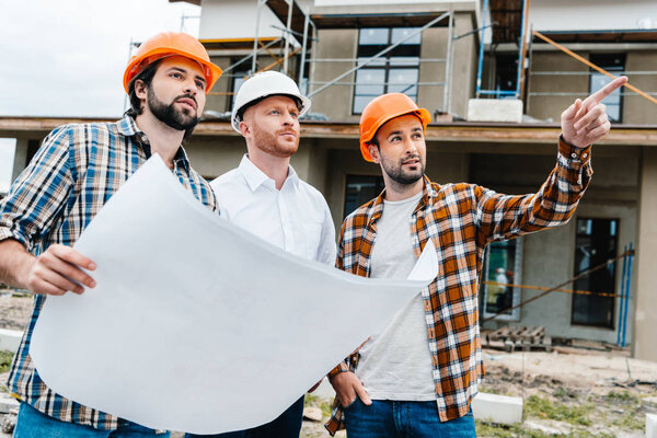 группа архитекторов с планом строительства, стоящих перед строительной площадкой и смотрящих в сторону
