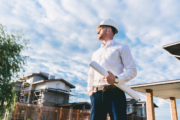 вид снизу красивого архитектора в белой рубашке с чертежом, стоящим во дворе перед домом и смотрящим в сторону

