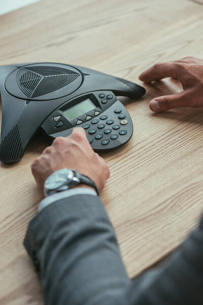 cropped shot of businessman pushing button of conference phone at workplace in office