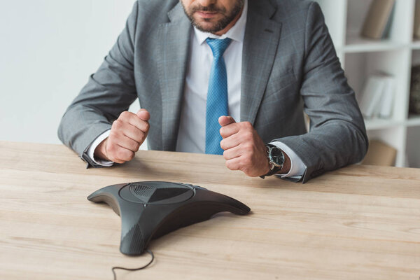 cropped shot of failed businessman sitting in front of conference phone and making fists at office
