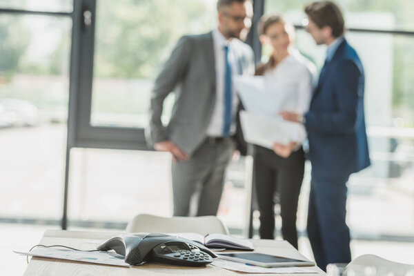 крупный план конференц-телефона с размытыми деловыми людьми на заднем плане в офисе
