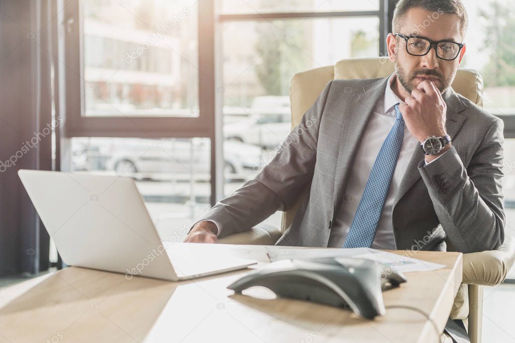 handsome thoughtful businessman sitting at workplace and looking away