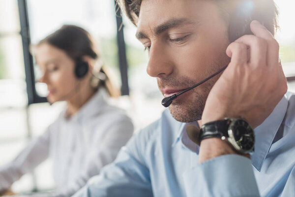 handsome call center manager in headphones with mike sitting at workplace while his colleague sitting on background