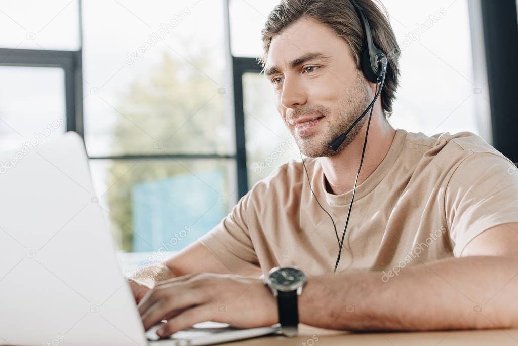 smiling young support hotline worker with laptop and headphones at workplace