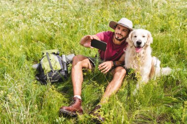 traveler in hat taking selfie on smartphone with dog on summer meadow clipart