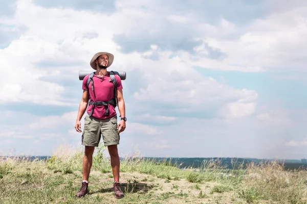 英俊的徒步旅行者与背包站立在夏天草甸与多云天空 — 图库照片