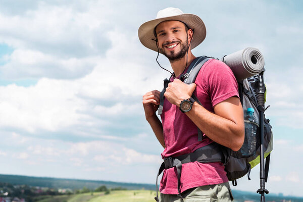 улыбающийся путешественник в шляпе с рюкзаком и туристический коврик, смотрящий в камеру
