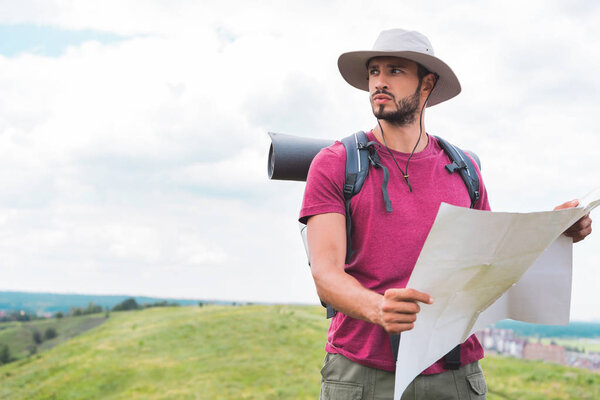 young traveler in hat with backpack holding map on summer meadow