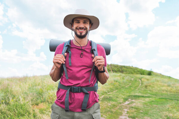 smiling hiker in hat with backpack walking on green field 