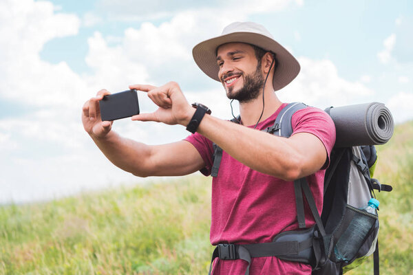 traveler with backpack taking photo on smartphone on summer meadow