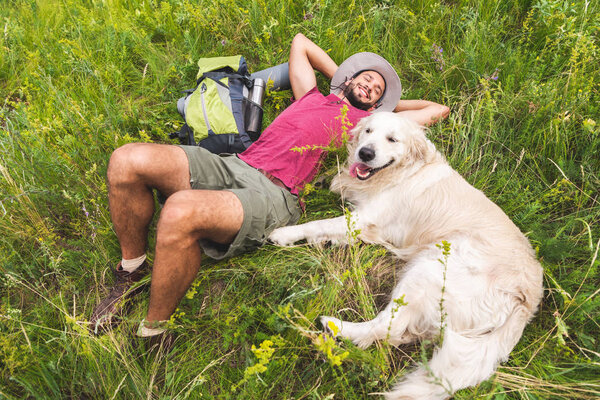 вид сверху улыбающегося путешественника и золотистого ретривера, лежащего на зеленой траве с рюкзаком

