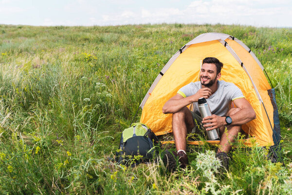 tourist with backpack and thermos sitting in yellow tent on summer meadow