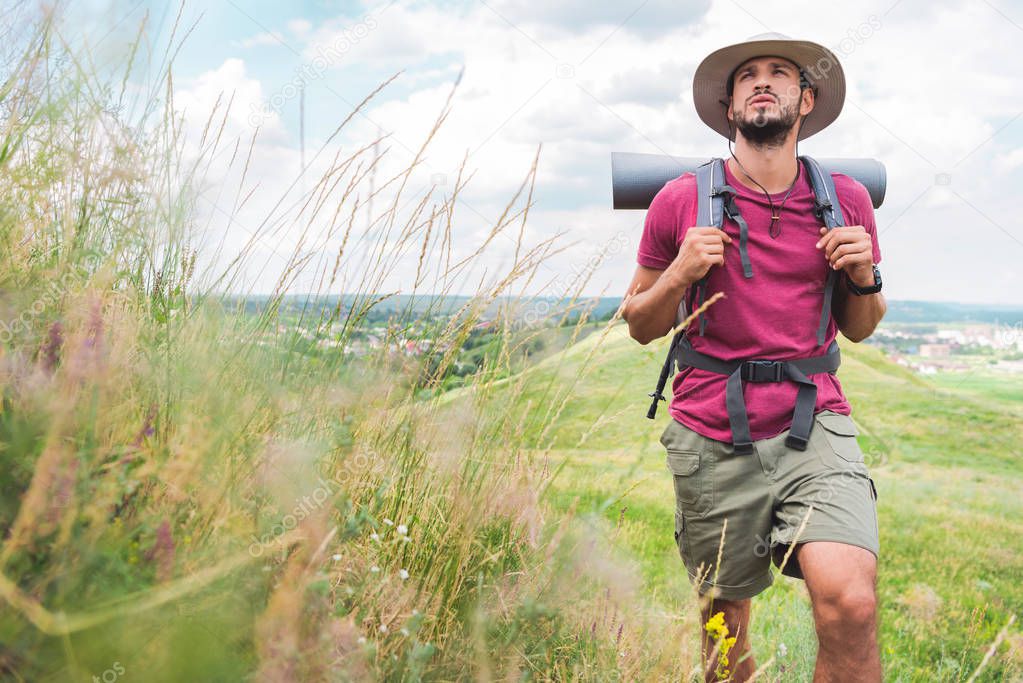 handsome hiker in hat with backpack and tourist mat walking on path on green meadow