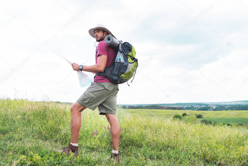 hiker with backpack holding map and standing on green field 
