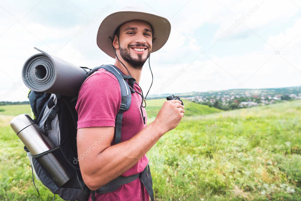 smiling traveler in hat with backpack holding binoculars on meadow