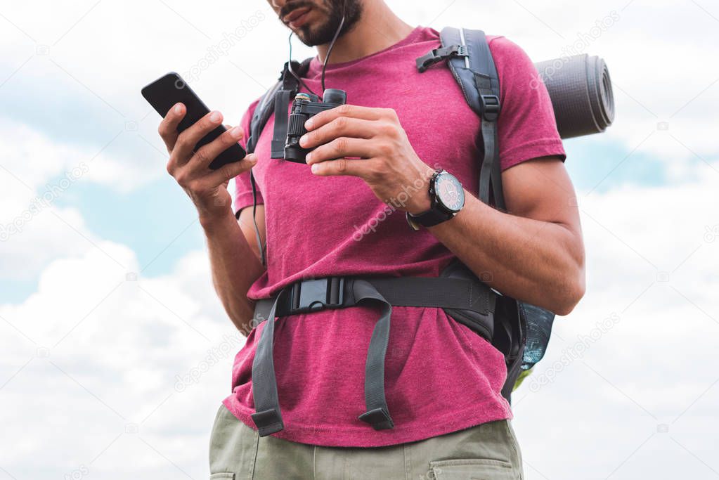 cropped view of tourist with backpack and binoculars using smartphone 