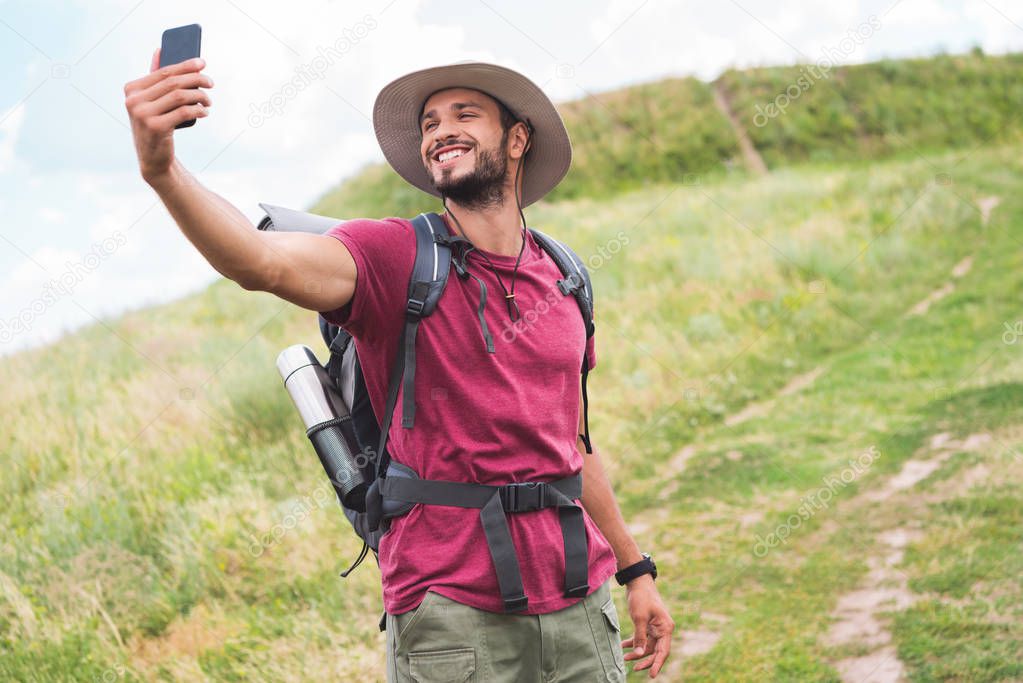 traveler with backpack taking selfie on smartphone on summer meadow