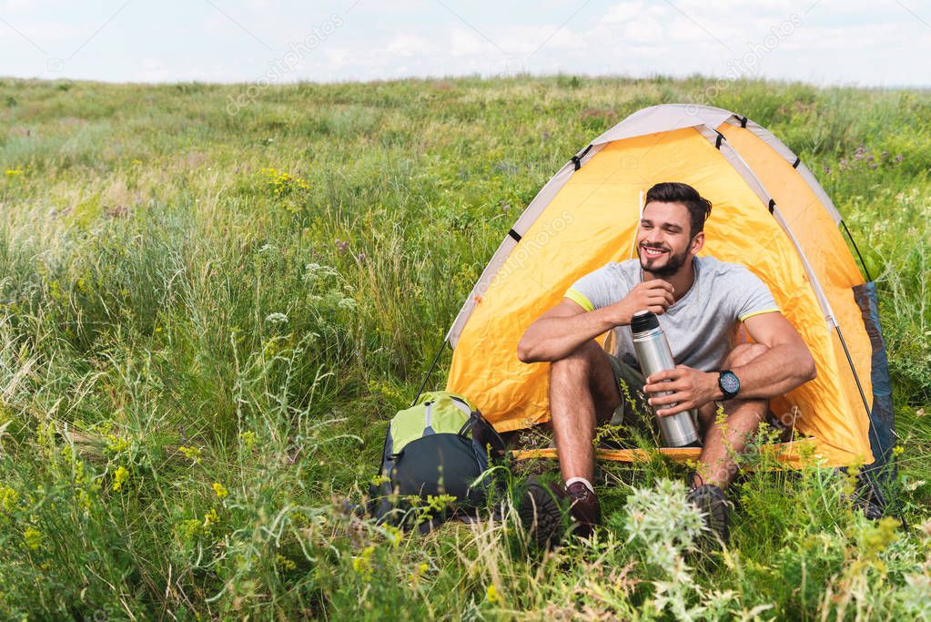 tourist with backpack and thermos sitting in yellow tent on summer meadow